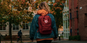 A young woman in coat with a red Kanken backpack walks through college campus in the autumn, back turned to us.
