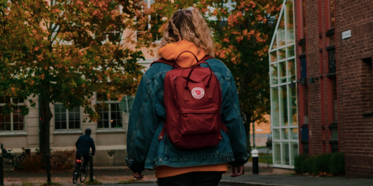 A young woman in coat with a red Kanken backpack walks through college campus in the autumn, back turned to us.