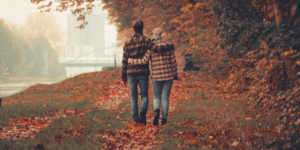 A couple walks together in an autumn park setting surrounded by fall foliage, viewed from behind.