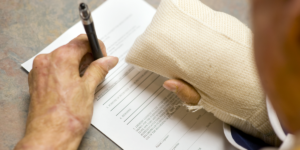 Close up of a person's hands on a counter, filling out a form. The right hand is completely bandaged, the left hand holds a pen.