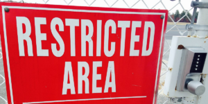 Red sign with white letters hanging on wire fence of a secure facility beside electronic key pad reads: RESTRICTED AREA