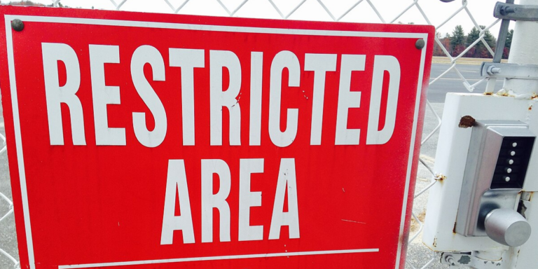Red sign with white letters hanging on wire fence of a secure facility beside electronic key pad reads: RESTRICTED AREA
