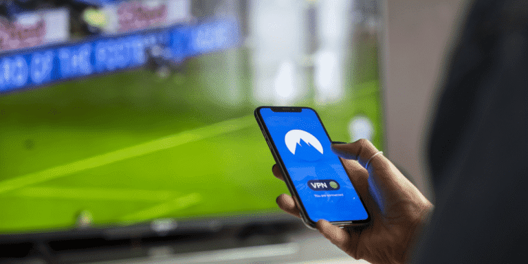 A person's hand holding a smartphone which displays a generic VPN program. In the background, a slightly blurry sports program is visible.